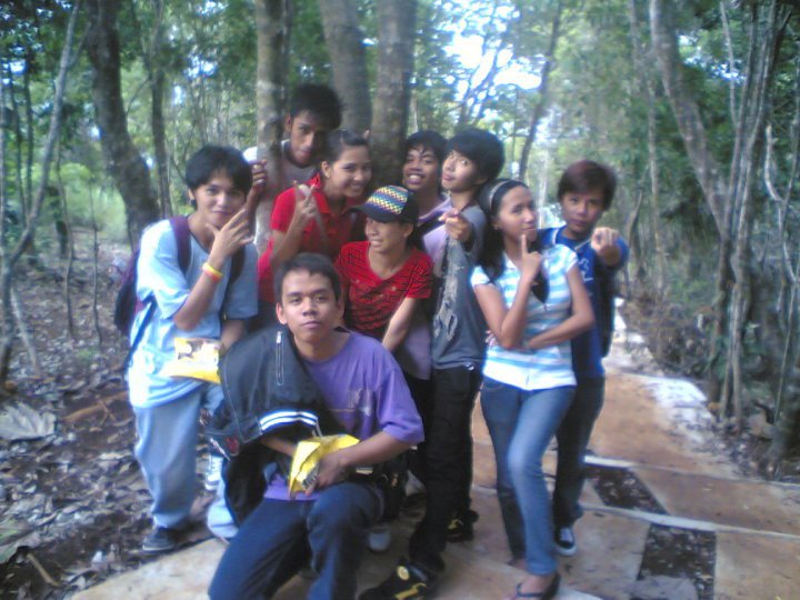 A group of young students posing together on a forest trail at Ermita Hill. They are smiling, doing playful hand signs, and standing close to each other among the trees.