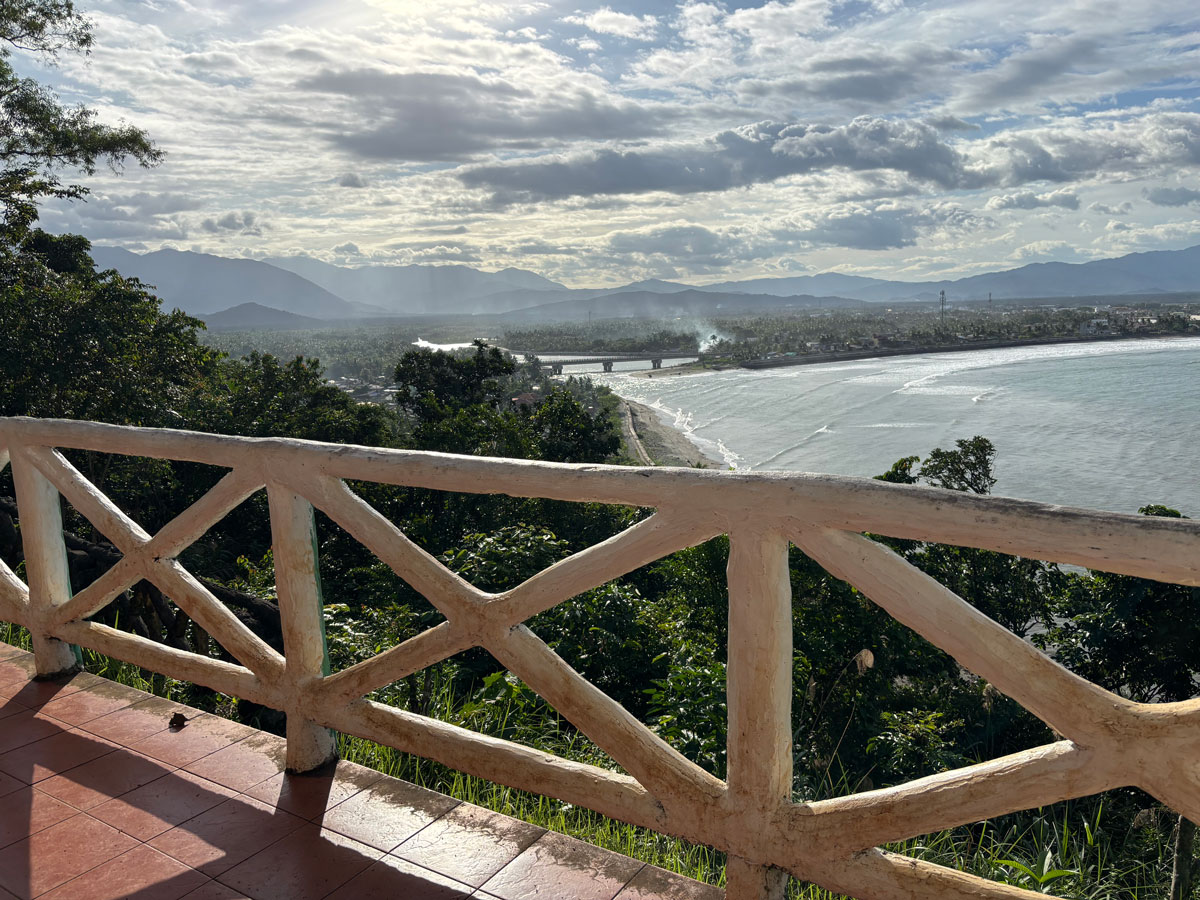 Scenic coastal view from Ermita Hill in Baler with mountain range, river bridge, lush greenery, and waves along the shoreline under a bright cloudy sky.