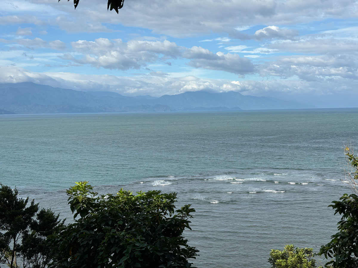 Wide ocean view from Ermita Hill in Baler, showing gentle waves, deep blue-green water, and a backdrop of distant mountain ranges beneath a partly cloudy sky.