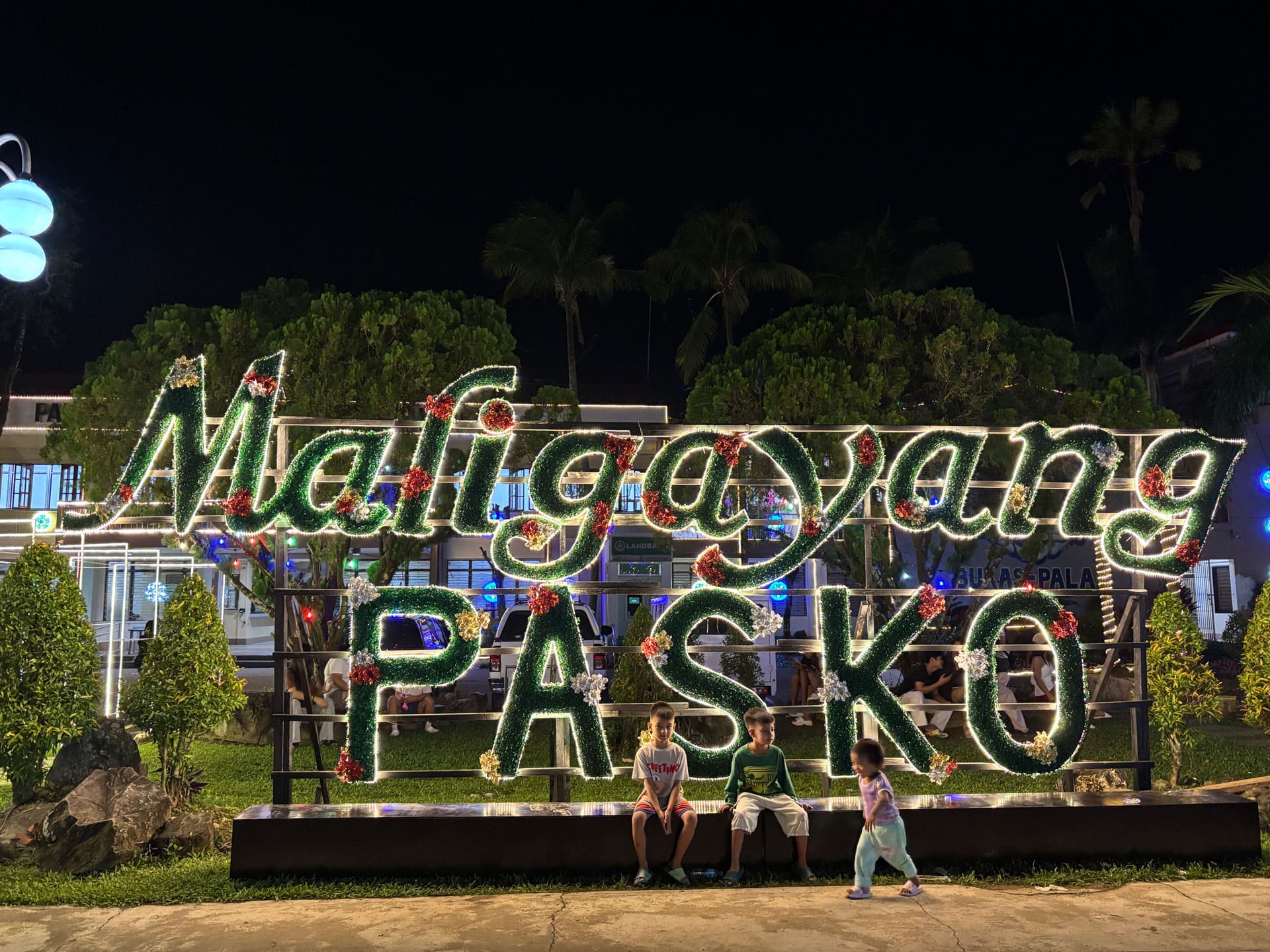 A large, illuminated green and red "Maligayang PASKO" sign at the Aurora Provincial Capitol grounds during PAILAW 2025, with children sitting and playing in front of the display.
