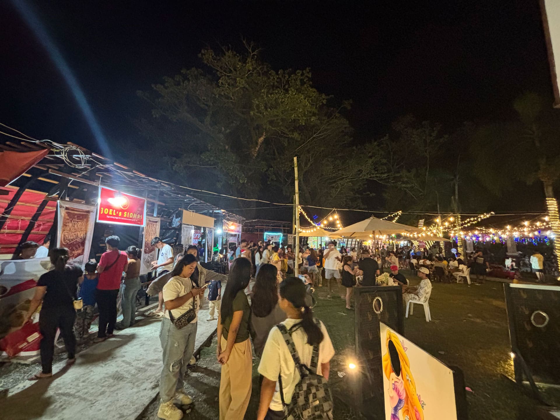 A busy night market scene at the Aurora Provincial Capitol grounds during PAILAW 2025, featuring food stalls like "Joel's Siomai" and people lining up for snacks under string lights.