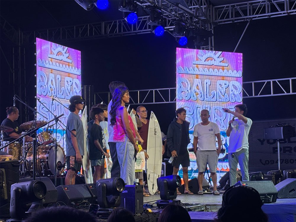 Awarding ceremony during BALER SURF FEST 2025. Several participants, organizers, and winners stand on a stage with surfboards while a host speaks into a microphone. Large LED screens in the background display "BALER SURF FEST," with stage lights illuminating the event and an audience watching from the front.