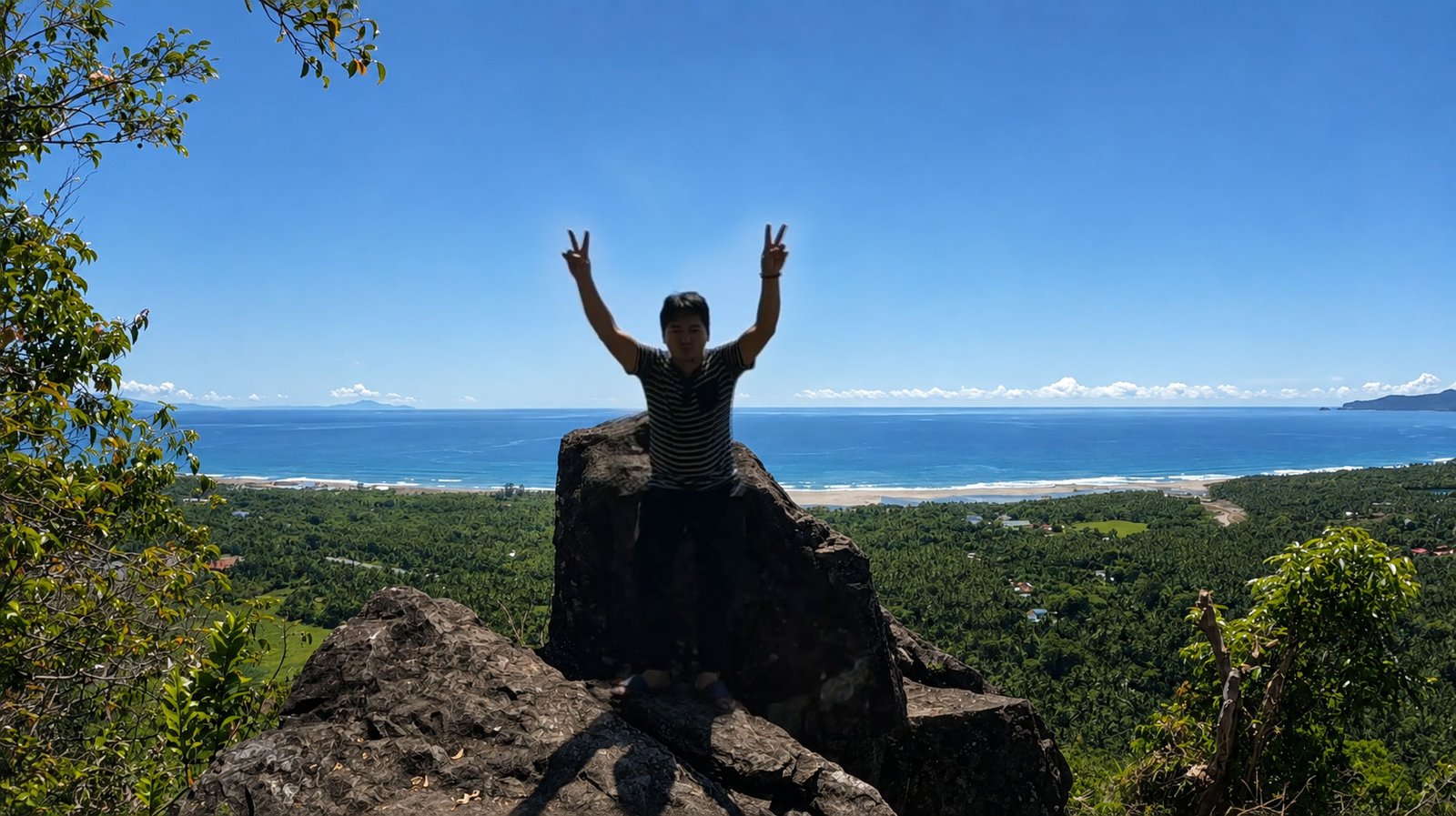 Traveler standing on the rocky summit of Bantay Lobbot overlooking lush green forests, a white sand coastline, and the blue ocean under a clear sunny sky in Dipaculao, Aurora, Philippines.